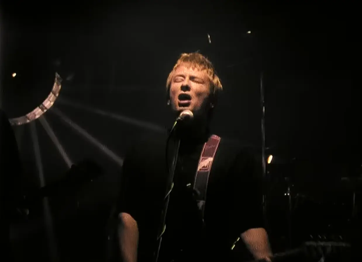 Thom Yorke performing 'Creep' live with closed eyes, singing emotionally into a microphone while playing electric guitar, under dramatic dark stage lighting with visible light beams in the background.